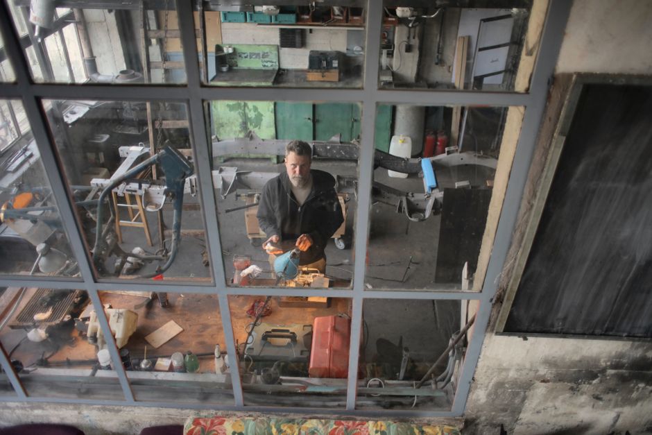 From above view of serious bearded male mechanic in casual wear standing near workbench with various equipment in garage and looking at camera